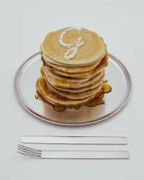 Tall stack of golden pancakes with maple syrup and powdered sugar letter on a metal plate with flatware