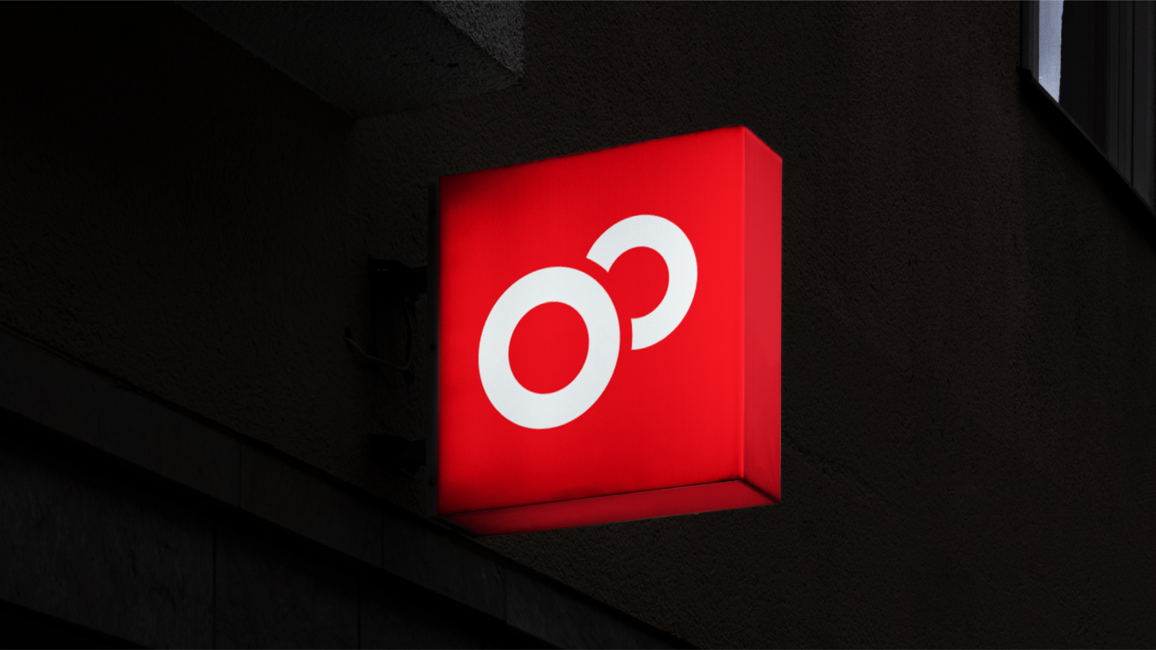 Illuminated red sign with a white graphic logo mounted on the side of a dark building during the night, creating a stark contrast.