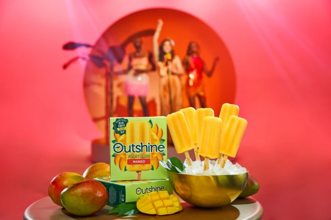 Mango ice popsicles and mangoes are displayed on a table with boxes of Outshine fruit bars in a brightly lit studio setting.