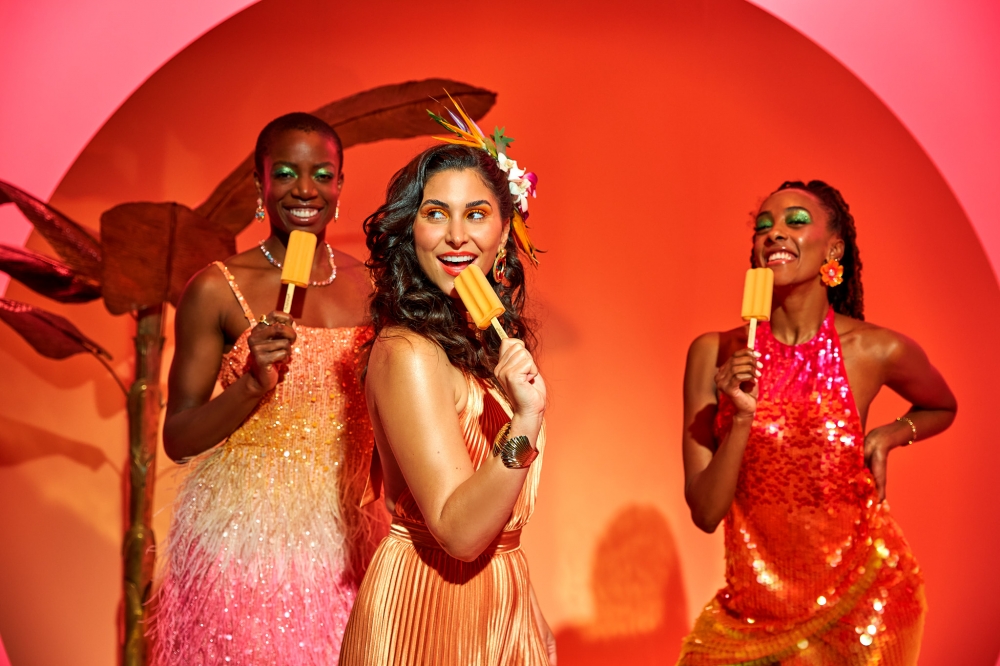 Three stylish women in sequined dresses are indoors against a vibrant orange background, each enjoying a colorful popsicle.