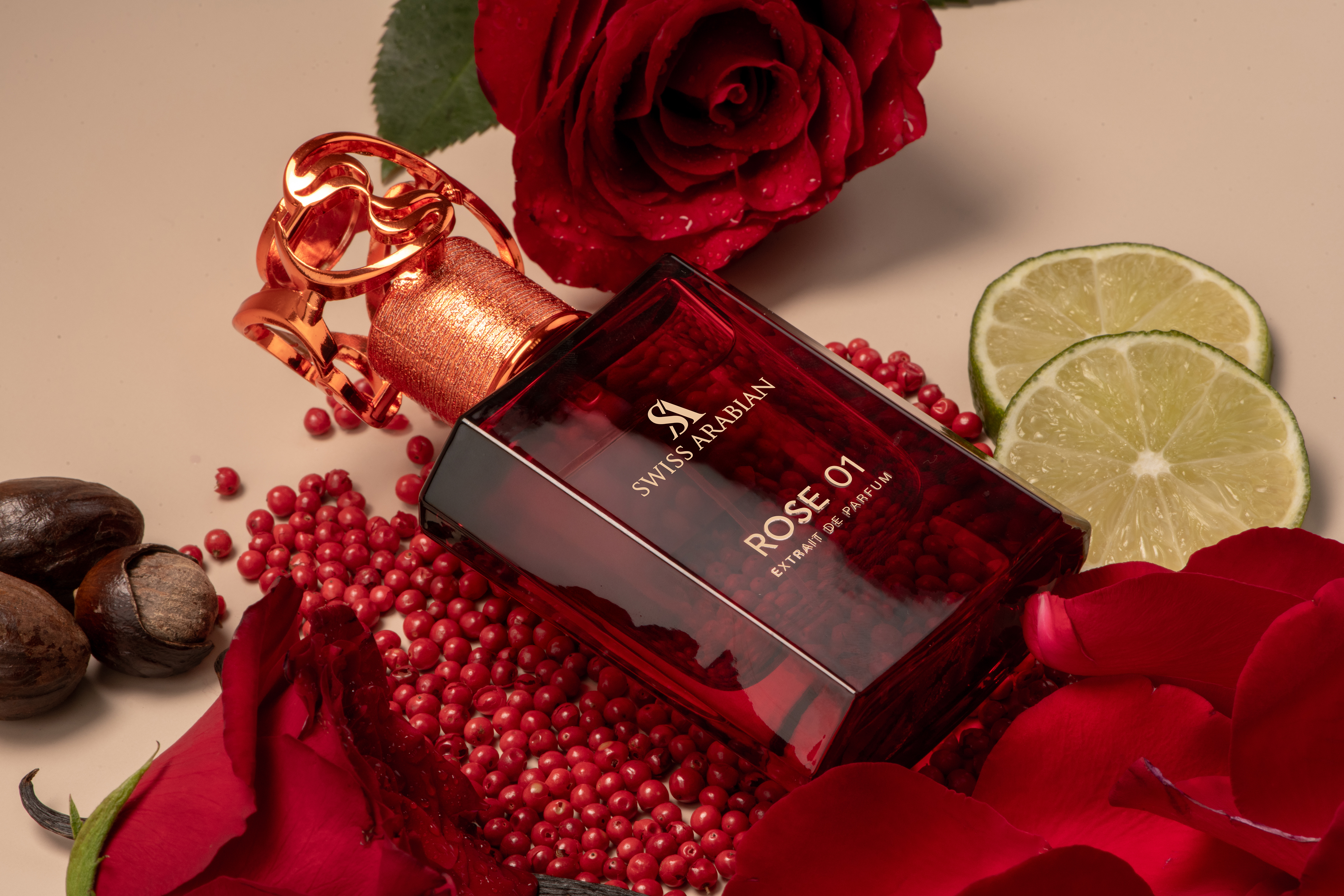Still life of Swiss Arabian Rose 01 perfume bottle surrounded by red rose petals, pink peppercorns, limes, and brown nuts on a beige surface.