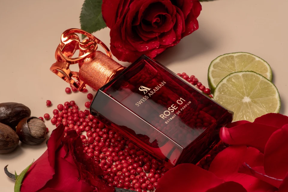 Still life of Swiss Arabian Rose 01 perfume bottle surrounded by red rose petals, pink peppercorns, limes, and brown nuts on a beige surface.
