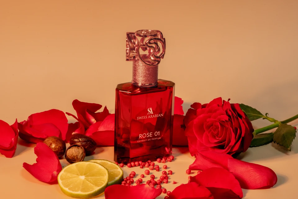 A vibrant studio shot features a red perfume bottle surrounded by red rose petals, lime slices, and peppercorns against a soft backdrop.
