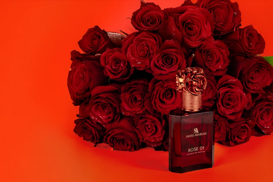 Dark red perfume bottle with gold cap, "Swiss Arabian" brand, surrounded by bunch of dark red roses on saturated red background.