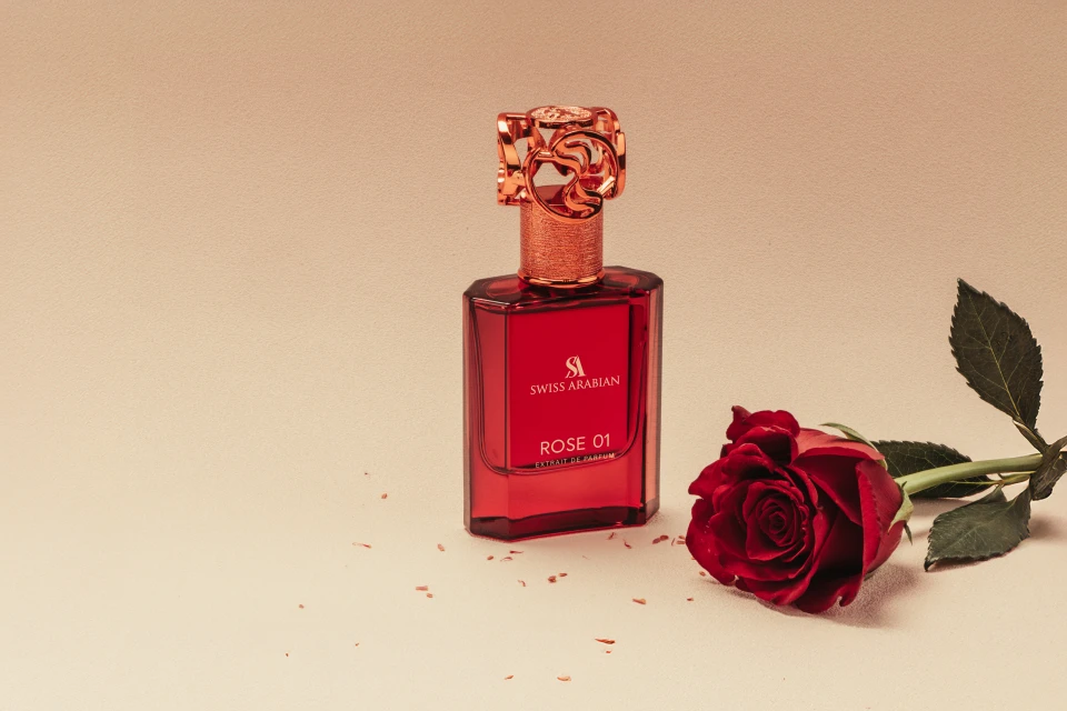 A red-toned still life featuring a 'Swiss Arabian' perfume bottle and a red rose on a cream-colored surface. Focus on beauty and fragrance.