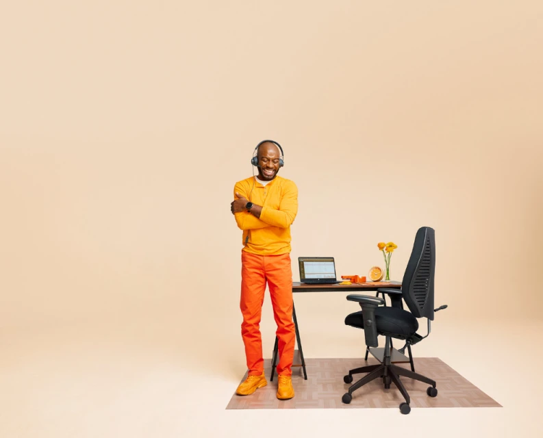 Smiling Black man in orange outfit stands near desk with laptop and black office chair in a neutral-toned indoor studio setting.