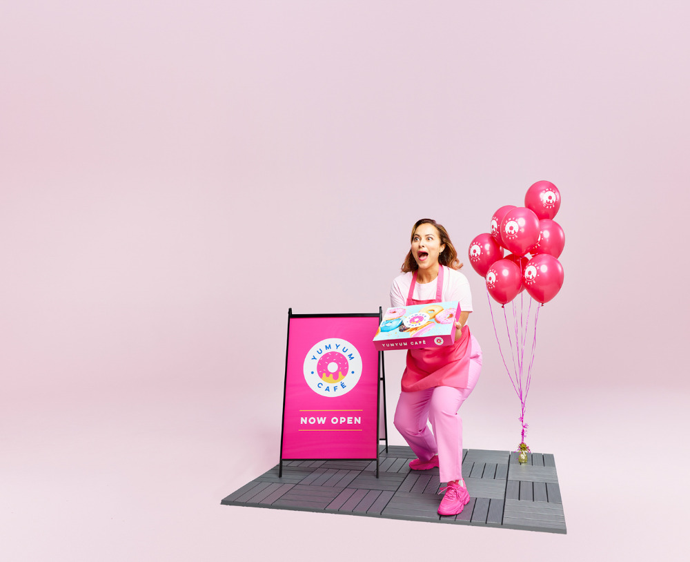 Enthusiastic woman in pink uniform, holding a donut box with balloons, promoting YumYum Cafe in a bright, eye-catching pink setting.
