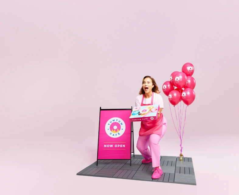 Enthusiastic woman in pink uniform, holding a donut box with balloons, promoting YumYum Cafe in a bright, eye-catching pink setting.