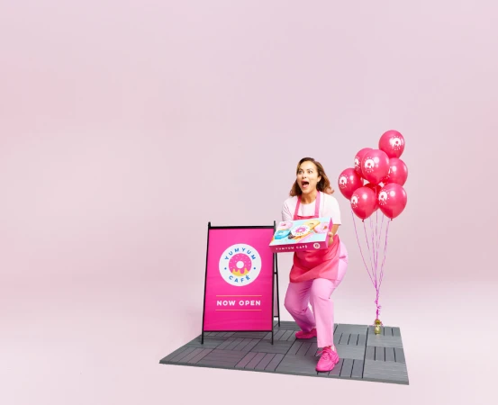 Enthusiastic woman in pink uniform, holding a donut box with balloons, promoting YumYum Cafe in a bright, eye-catching pink setting.