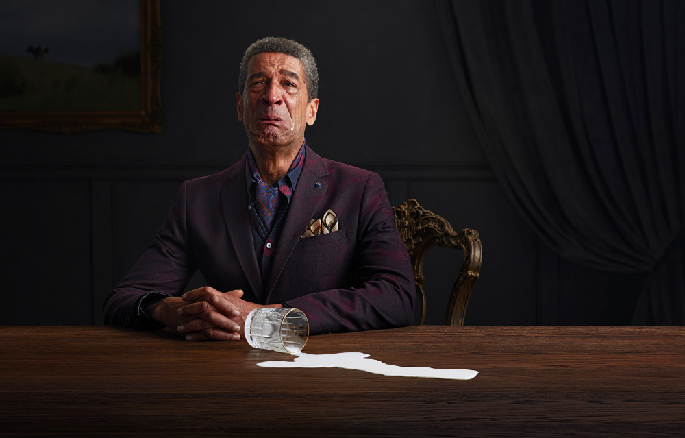 Mature man in a suit expressing sadness at a spilled drink on a wooden table in an indoor setting with dark background.