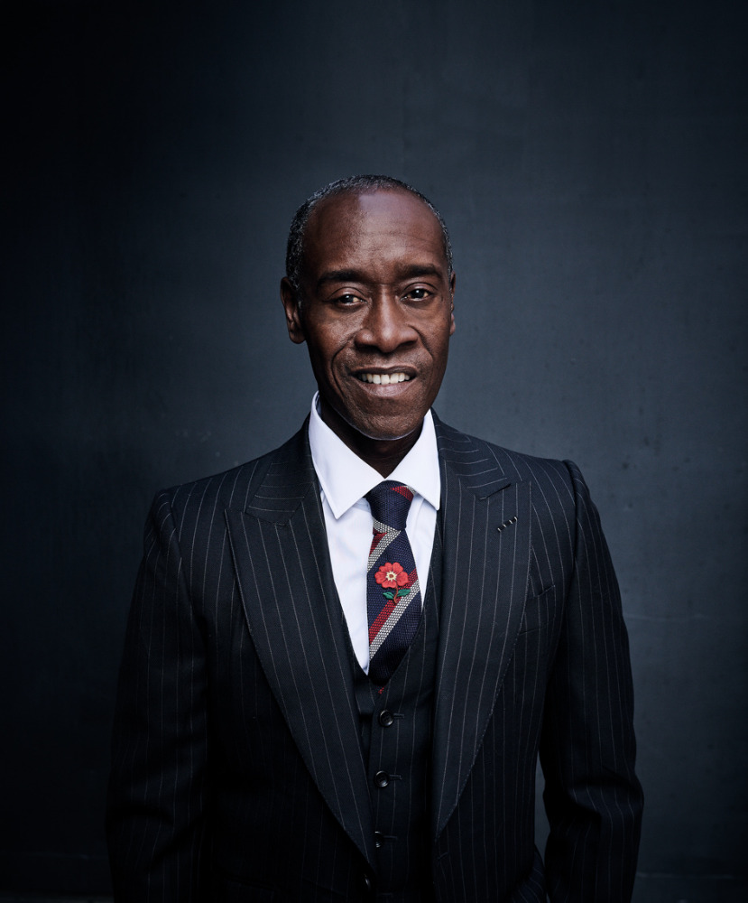 A portrait of Don Cheadle wearing a pinstripe suit, vest, and tie with a floral pin, smiling in front of a dark gray background.