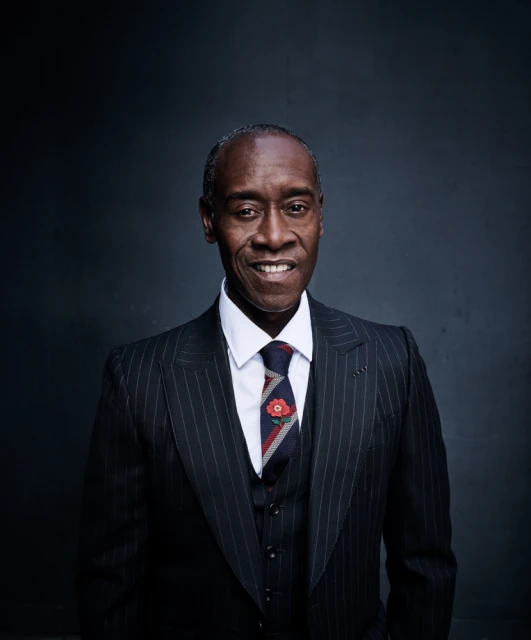 A portrait of Don Cheadle wearing a pinstripe suit, vest, and tie with a floral pin, smiling in front of a dark gray background.