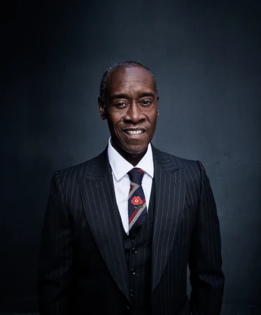 A portrait of Don Cheadle wearing a pinstripe suit, vest, and tie with a floral pin, smiling in front of a dark gray background.