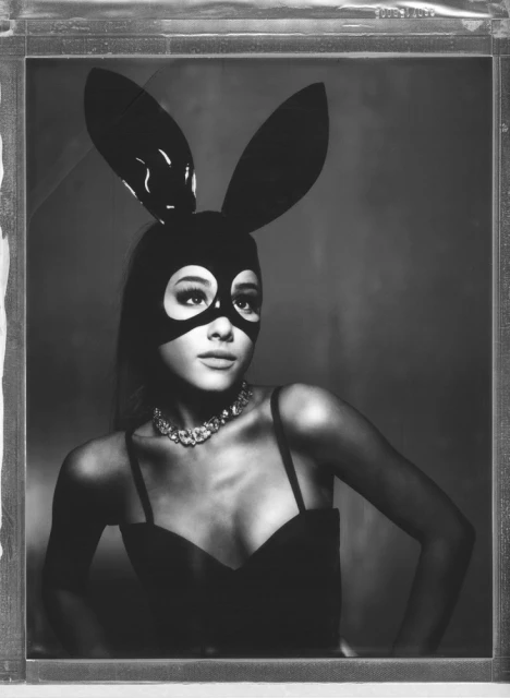 Monochrome portrait of a woman wearing a black bunny mask, jewelry, and strappy top, looking to the right, framed in a glass case.