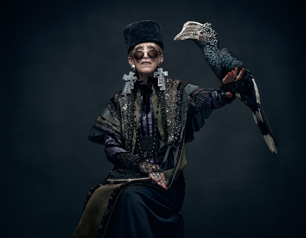 An elderly woman in ornate clothing poses with a black-and-white bird on her arm in a formal studio portrait against a dark background.
