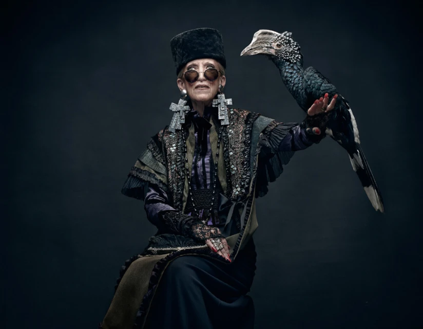 An elderly woman in ornate clothing poses with a black-and-white bird on her arm in a formal studio portrait against a dark background.