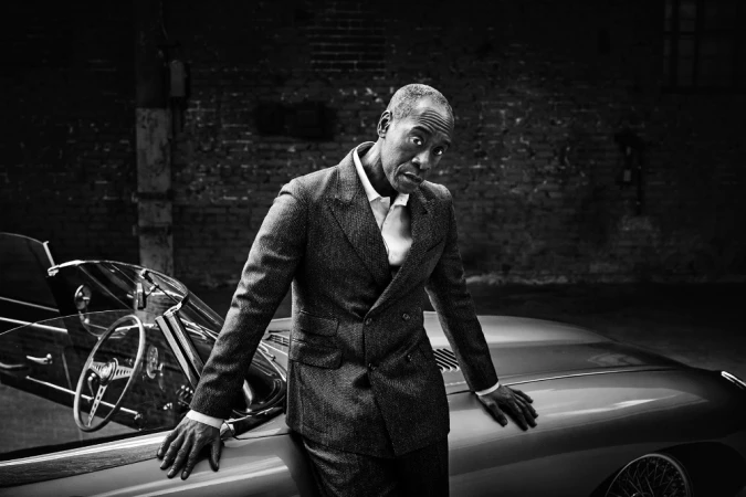 Adult man in a suit stands next to a vintage car in a black and white portrait, showcasing classic automotive style and fashion.