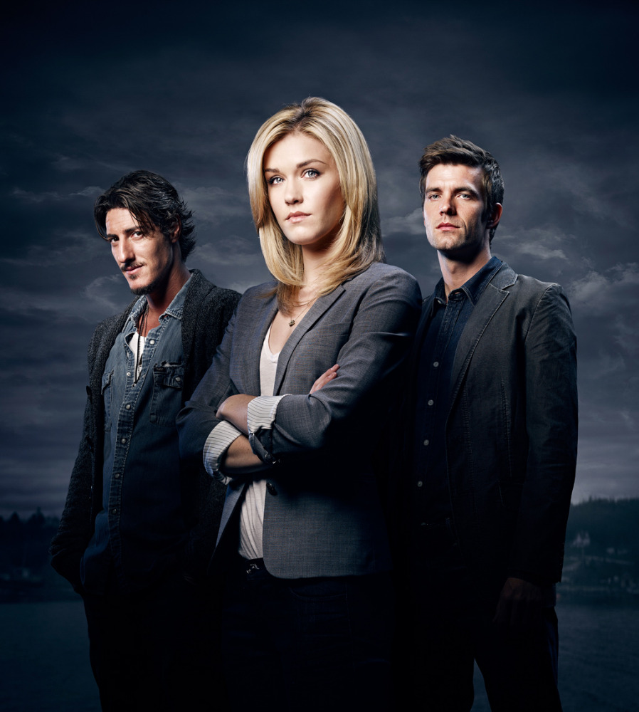 Studio portrait of three actors in blazers and a denim jacket against a cloudy dark blue sky with water and land visible in the distance.