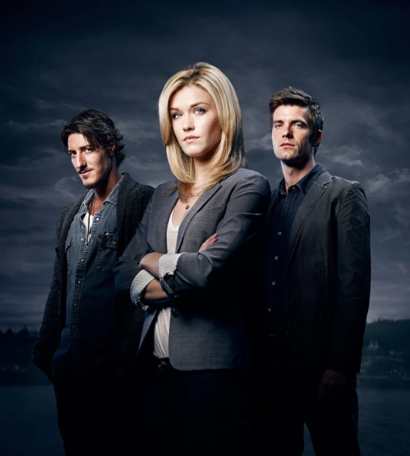 Studio portrait of three actors in blazers and a denim jacket against a cloudy dark blue sky with water and land visible in the distance.