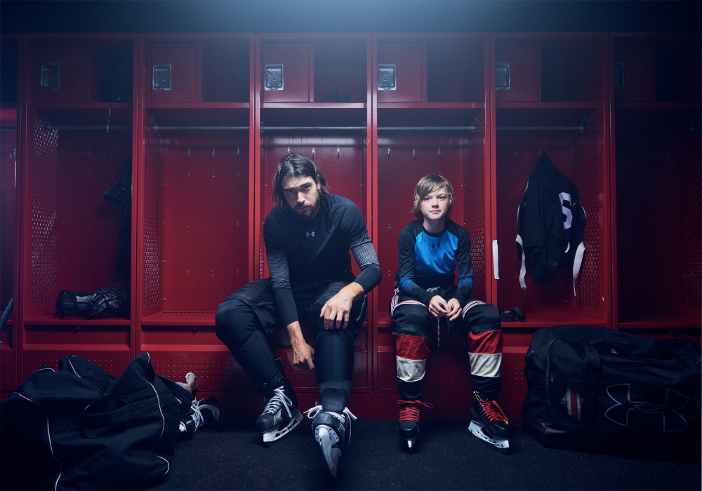 An adult and child hockey player are shown seated in a locker room, both in full gear and ready for the game in a medium indoor shot.