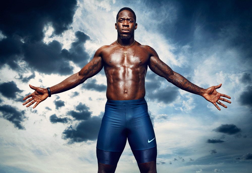 Muscular African American man with arms outstretched wearing athletic shorts posing in front of a dramatic blue and white sky background.