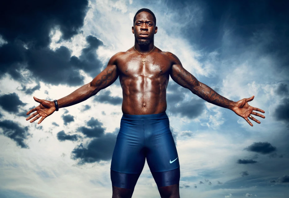 Muscular African American man with arms outstretched wearing athletic shorts posing in front of a dramatic blue and white sky background.