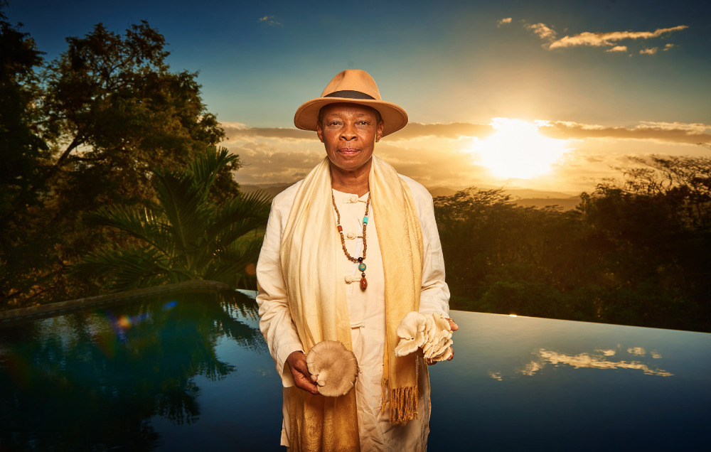 An older adult poses outdoors at dawn near an infinity pool and lush vegetation wearing a hat, necklace, and holding mushrooms.