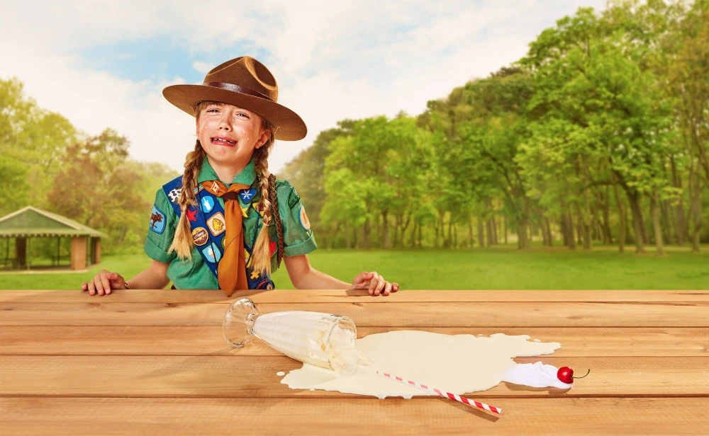 Crying girl in a scout uniform stands at a picnic table with a spilled milkshake, featuring a park, trees, and clear blue sky.