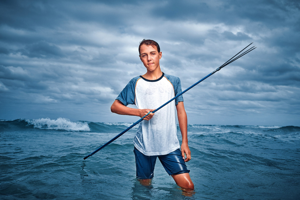 Young man in ocean stands waist-deep holding a spear, waves crash, and a cloudy sky fills the background, creating a striking image.