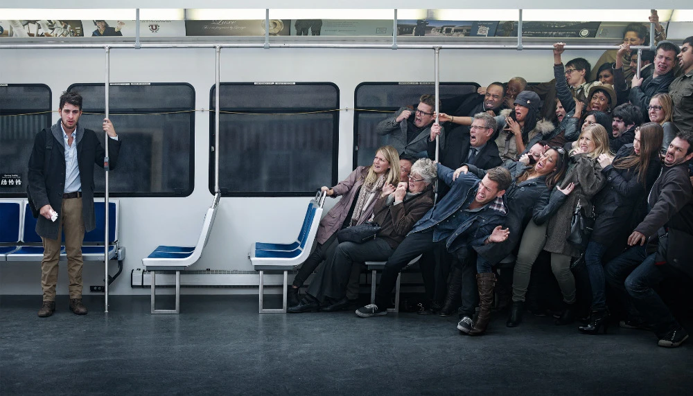 A man stands relaxed on a subway train, contrasting with a large group of passengers who are pressed together and yelling in the background.