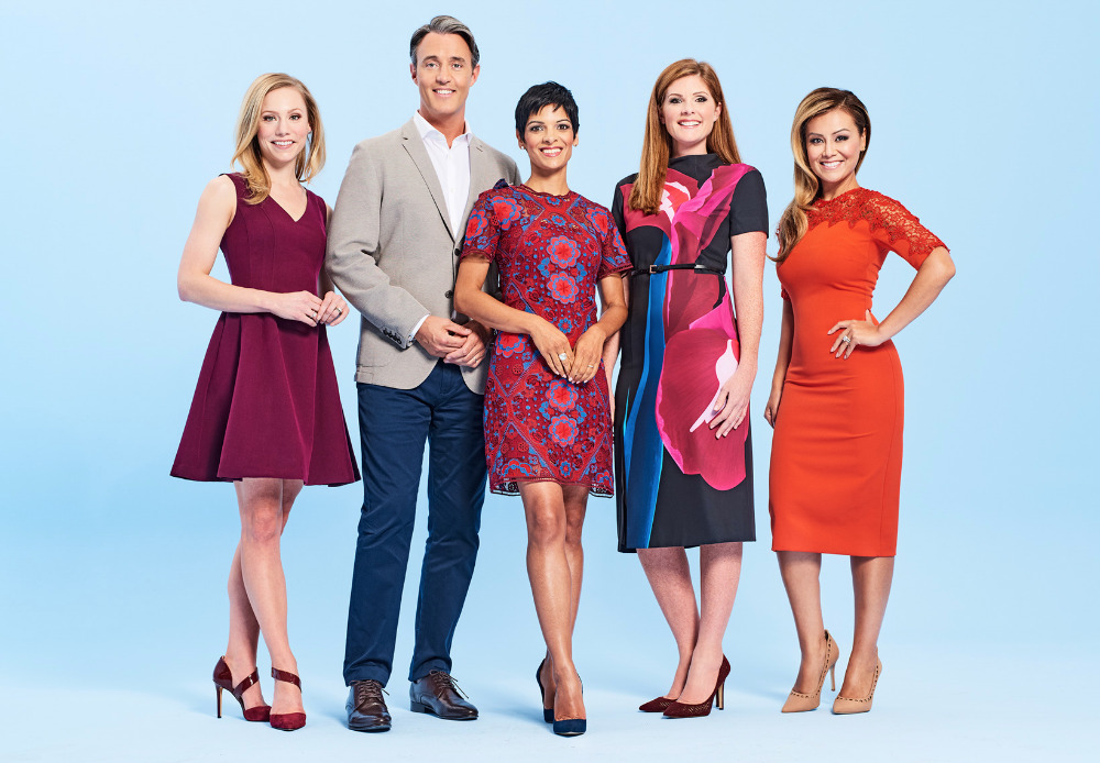 Group portrait of five news presenters in dresses and a jacket, standing in a row against a studio backdrop with a light blue gradient.