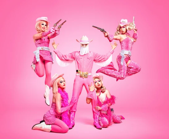 Group of five drag queens in pink Western wear posing with toy guns against a pink background in a studio setting.