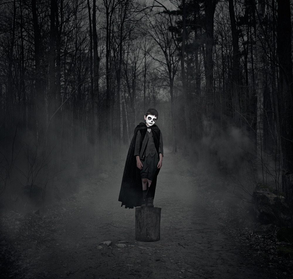 Child in Halloween costume with skull face paint standing on a tree stump in a dark, misty forest wearing a cape.