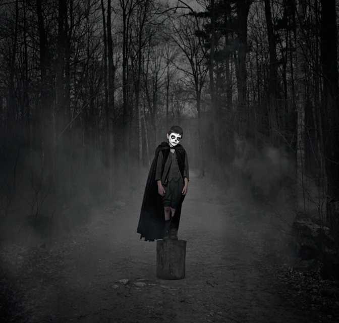 Child in Halloween costume with skull face paint standing on a tree stump in a dark, misty forest wearing a cape.