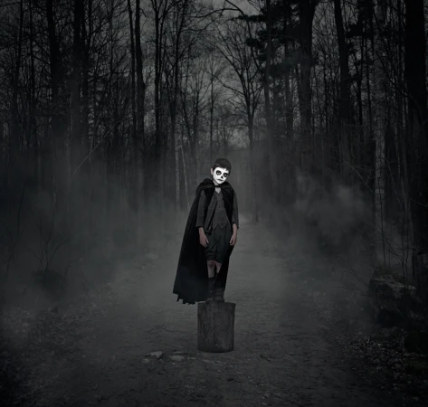 Child in Halloween costume with skull face paint standing on a tree stump in a dark, misty forest wearing a cape.