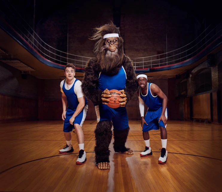 A basketball team photo with a large hairy mascot in blue and two male basketball players in a gymnasium with a wooden court.