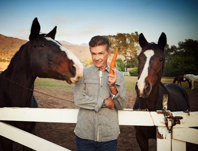 Man with two horses outside; he is holding a carrot in his hand. Landscape background shows fields, trees, and a house.