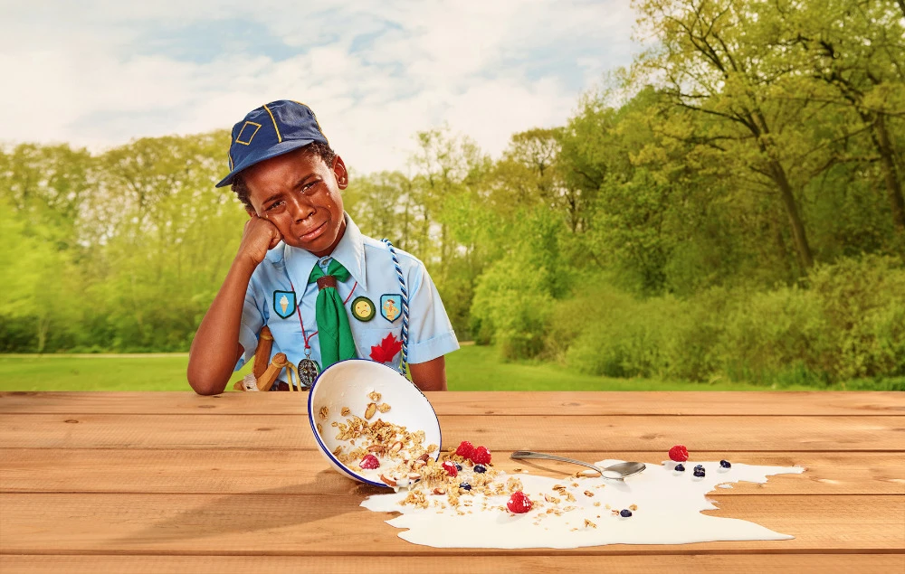 A young boy in a scout uniform sits at a picnic table with an upset facial expression next to a spilled bowl of cereal and berries.