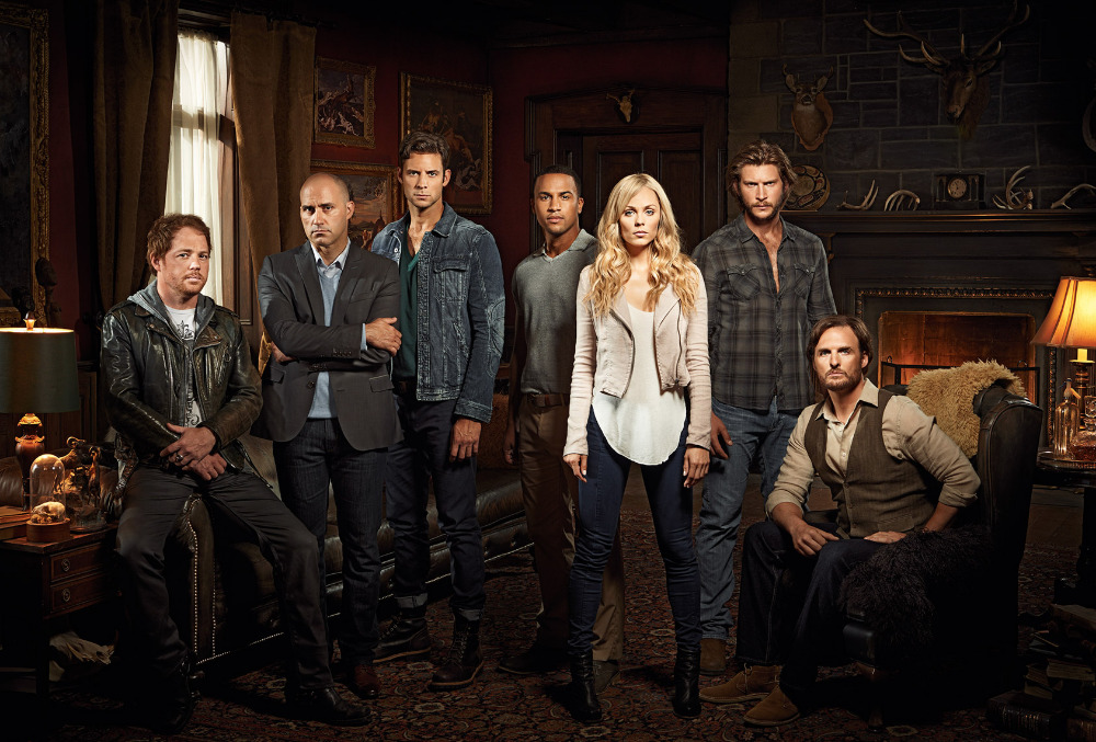 Group portrait of seven actors in casual clothing posing in a dark room with leather furniture and animal skulls mounted on the walls.