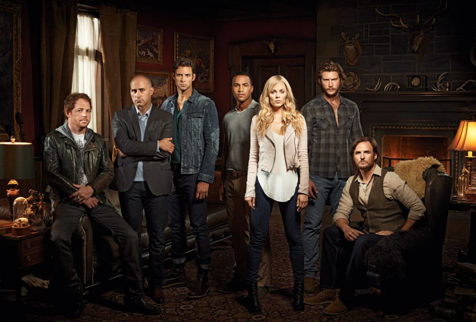 Group portrait of seven actors in casual clothing posing in a dark room with leather furniture and animal skulls mounted on the walls.