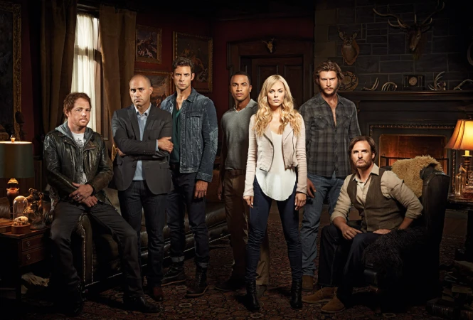 Group portrait of seven actors in casual clothing posing in a dark room with leather furniture and animal skulls mounted on the walls.