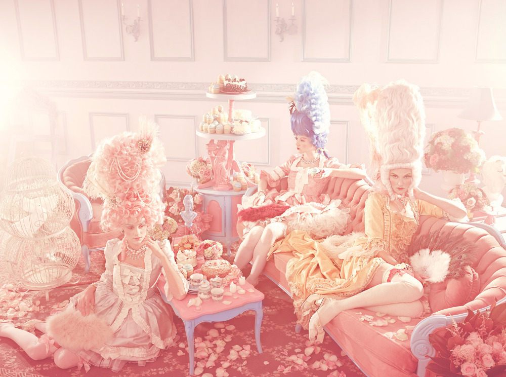 Three women in Marie Antoinette-inspired dresses and wigs lounge on pink furniture surrounded by flowers and pastries in an elegant interior.
