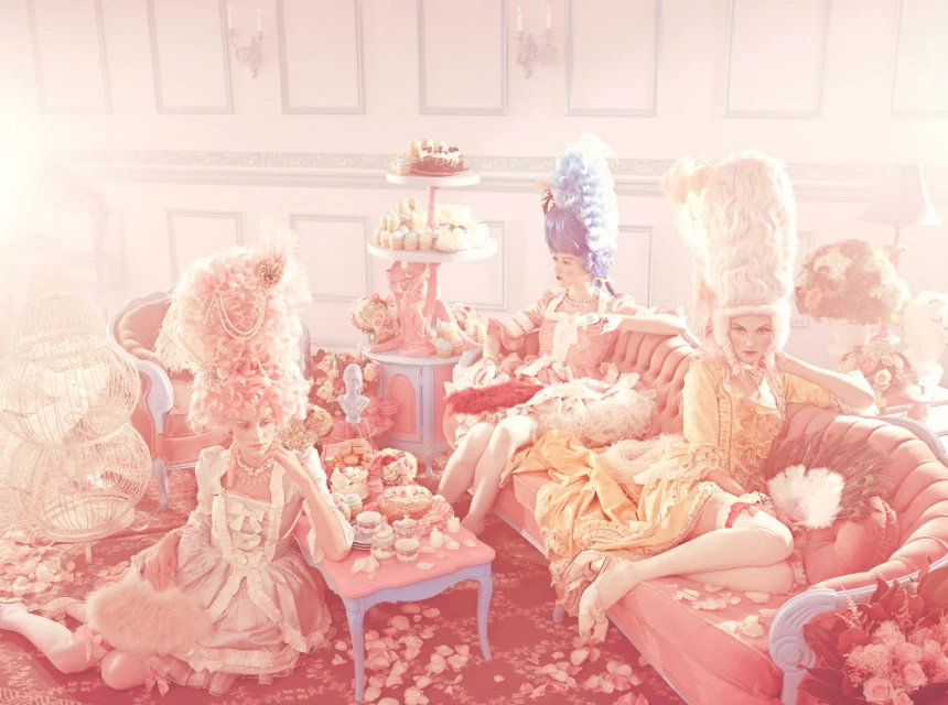 Three women in Marie Antoinette-inspired dresses and wigs lounge on pink furniture surrounded by flowers and pastries in an elegant interior.