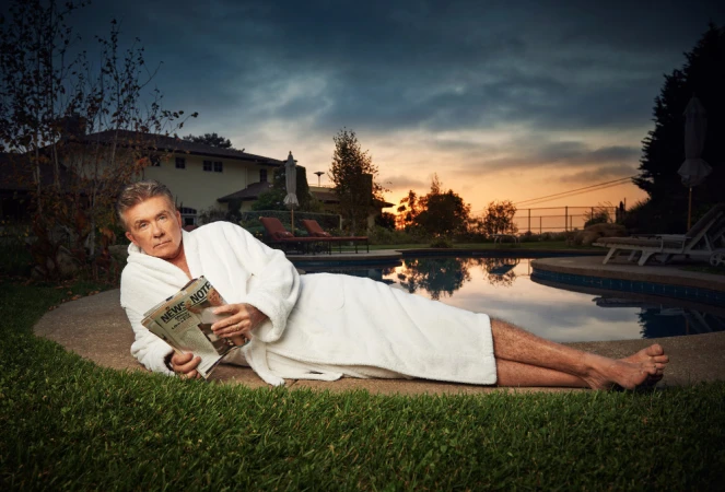 Mature man in white robe reclines on lawn with a newspaper, a pool and home in the background, under a cloudy sky at dusk.