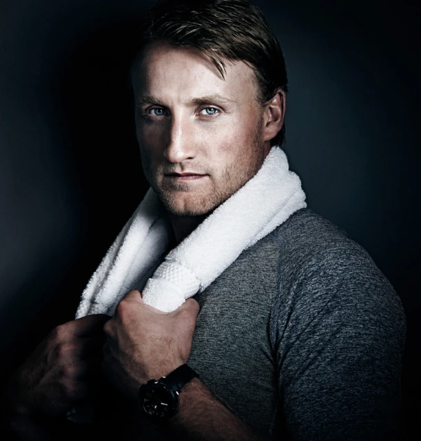 Portrait of a blonde adult male with a white towel draped over his shoulders, wearing a gray athletic shirt and a wristwatch, in a dark indoor setting.