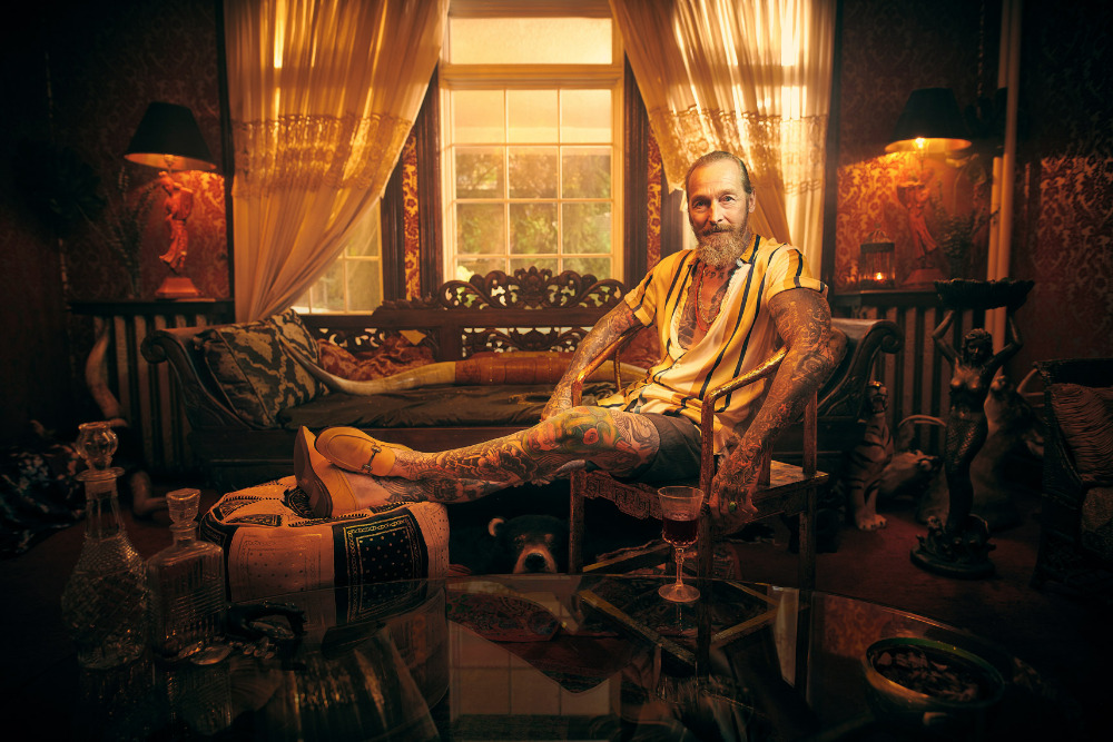 Tattooed adult male sitting in a chair in a heavily decorated room, wearing yellow and white striped shirt, with ornate furnishings.