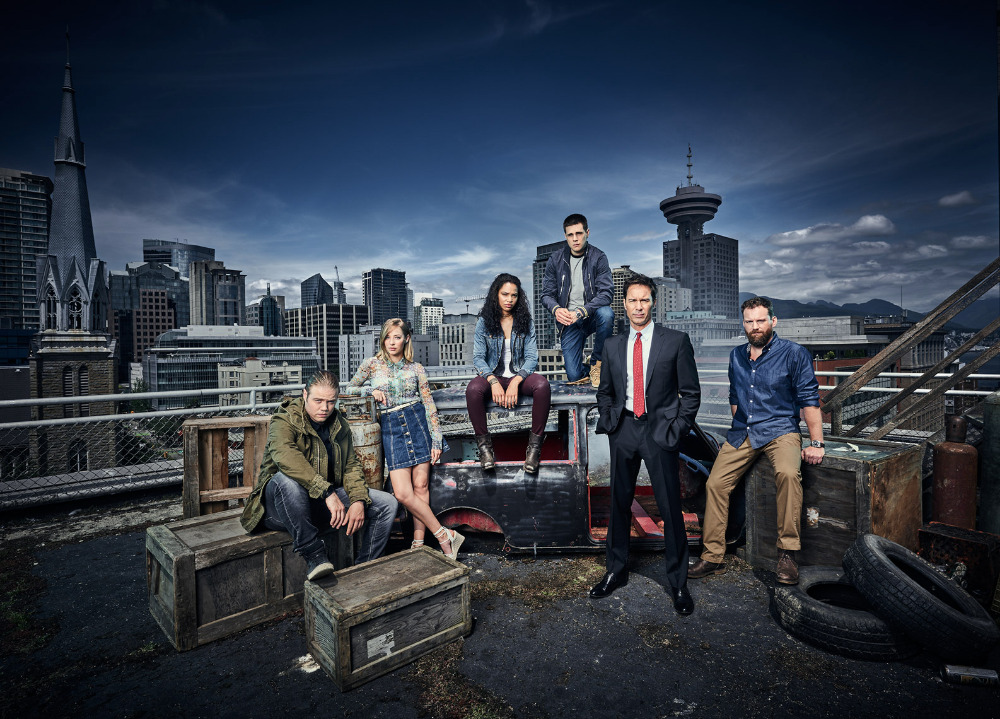 Group portrait of six people on a rooftop with a cityscape backdrop at night. Dramatic lighting and urban fashion create a cinematic mood.