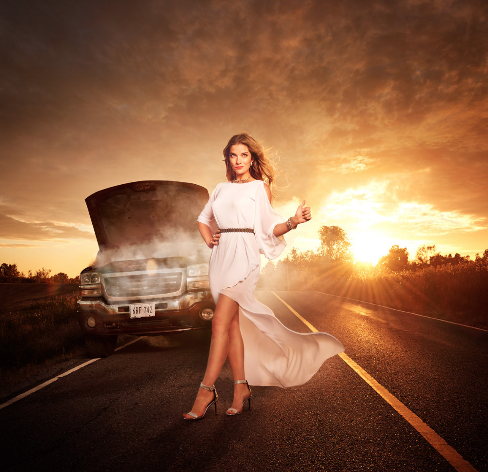 Woman in white dress hitchhiking on a road next to a smoking broken-down car with a sunset background. Automotive industry.