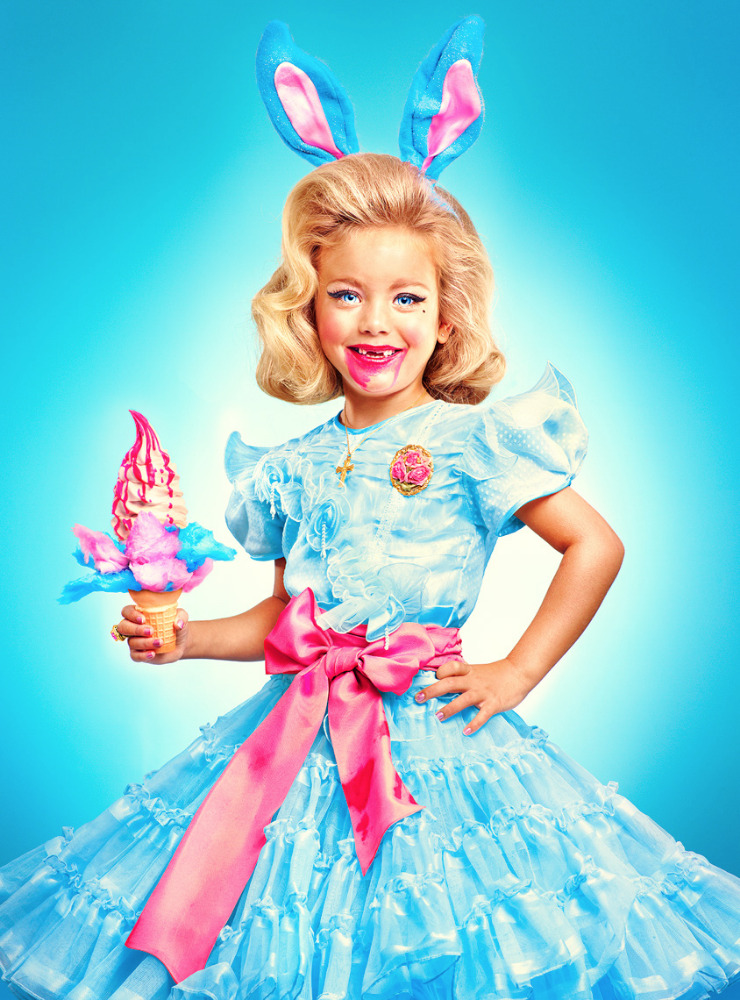 A young girl in a blue dress and bunny ears holds an ice cream cone, with bright, colorful makeup on a vibrant blue background.