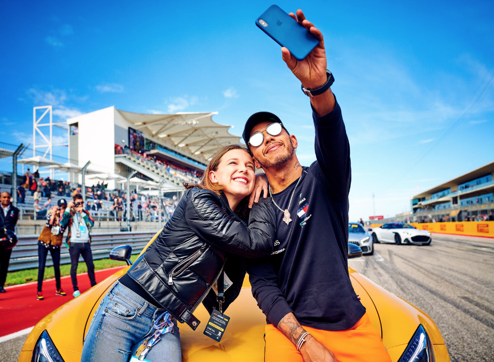 Millie Bobby Brown and Lewis Hamilton take a selfie while posing on a yellow sports car at an outdoor racetrack on a sunny day.
